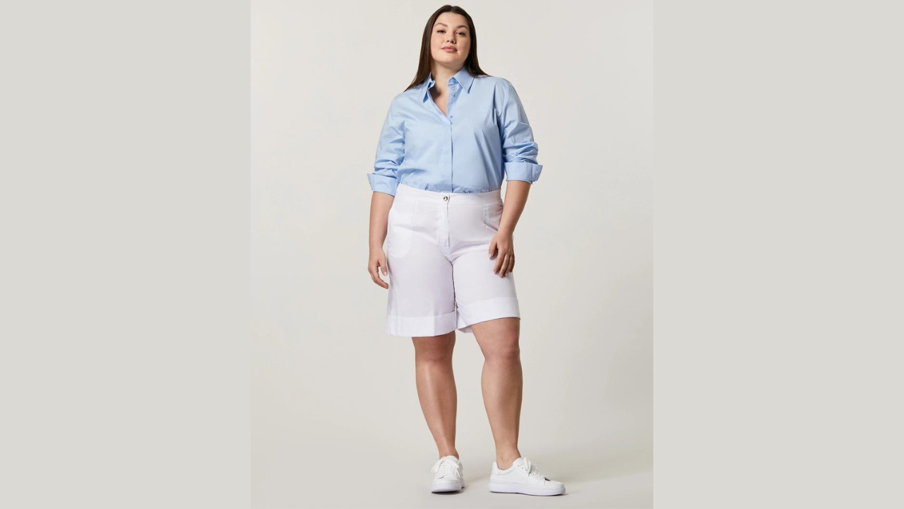 A picture of a woman wearing a blue shirt and white shorts, along with white sneakers, striking a pose for the camera. The theme is about how to beat the heat and dress with ease in the office 