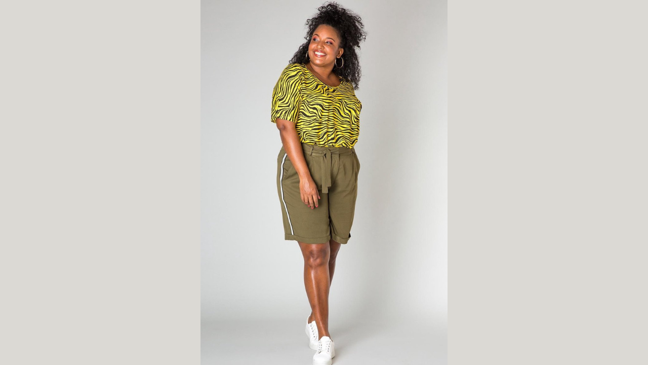 A photo of a black woman in a patterned t-shirt and green shorts, smiling and posing for the camera. The theme is about how to beat the heat and dress with ease in the office