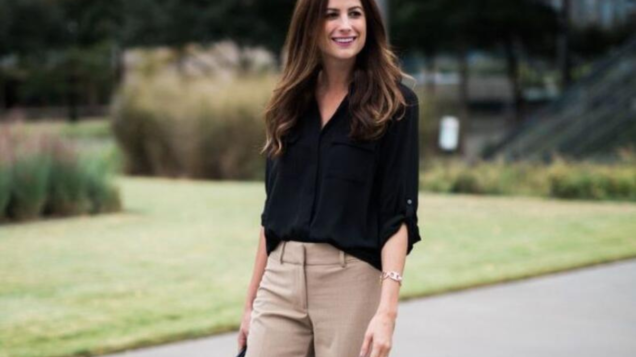 A picture of a woman with long hair, wearing a black shirt and brown pants, smiling as she walks. The theme is about how to beat the heat and dress with ease in the office