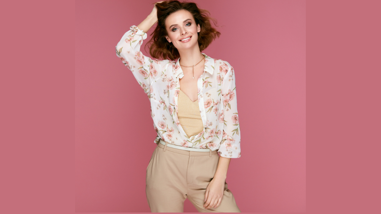 A picture of a woman with short hair, wearing a white blouse with a floral pattern and light-colored pants, striking a pose for the camera. The theme is about how to beat the heat and dress with ease in the office