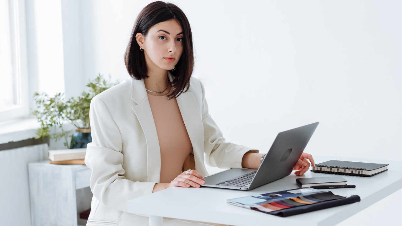 A photo of a woman with short hair, wearing a white blazer, sitting with a laptop and looking straight ahead.