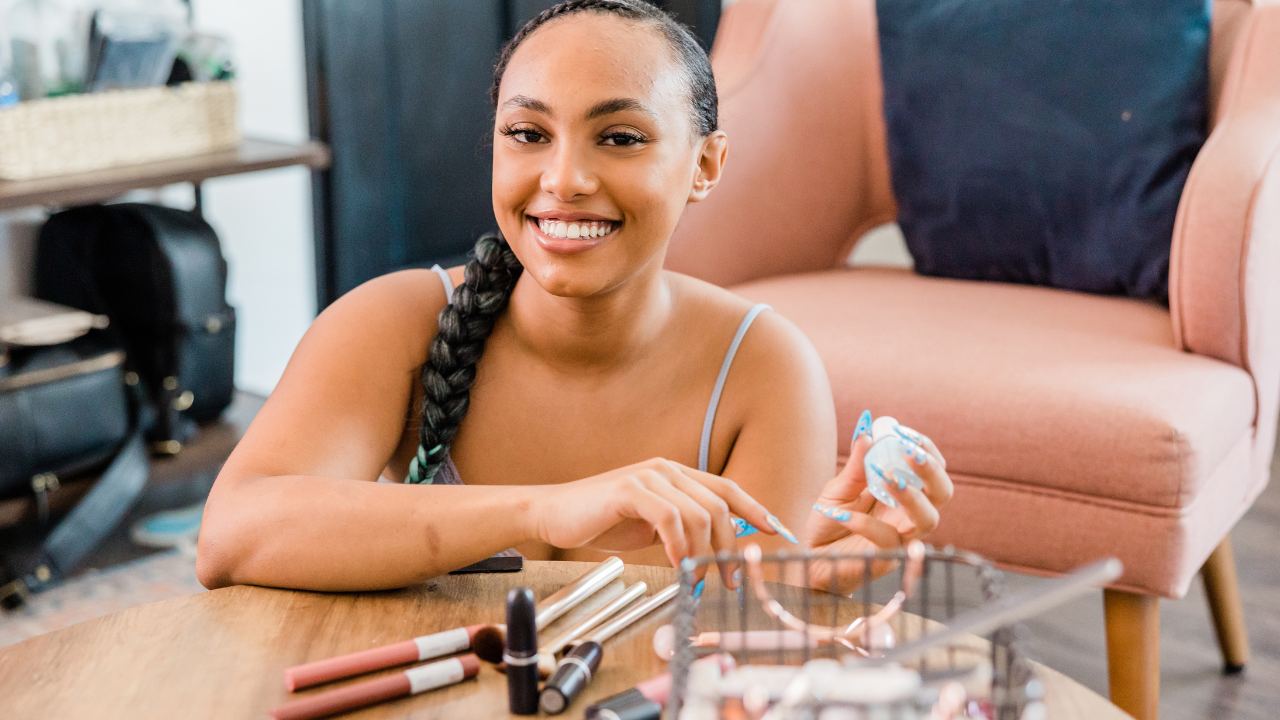A visual of a woman with a joyful expression, holding a makeup product, with various makeup items arranged on the table in front of her.