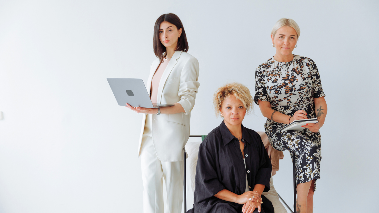 A visual featuring three women elegantly dressed in professional attire, posing for the camera. One is holding a laptop, another is clutching a notepad, and the third is seated gracefully.